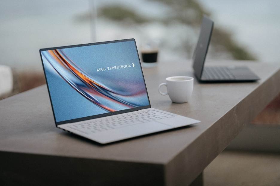 The image shows a sleek ASUS ExpertBook laptop placed on a wooden surface, with a cup of coffee on the desk and a clear view of the ocean in the background.
AI-generated content may be incorrect.